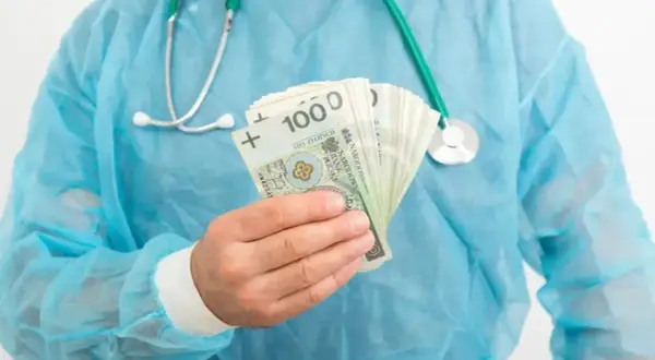 Lekarz rezydent w niebieskim fartuchu trzyma w dłoni plik banknotów 100 zł. Pytanie brzmi: ile zarabia lekarz rezydent?